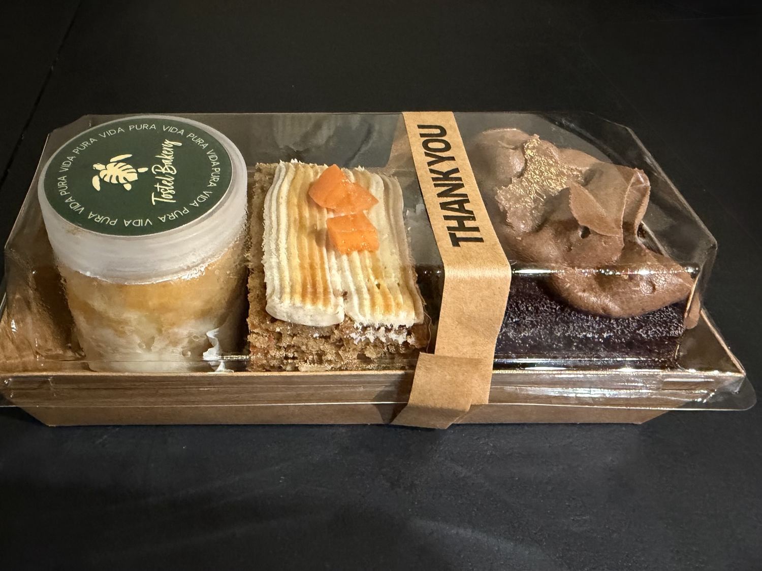 A Tostel gift box holding a tres leches jar, a carrot cake slice, and a chocolate cake, sealed with a Pura Vida label
